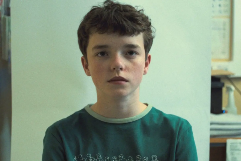 Foto da série Adolescência mostrando um adolescente olhando diretamente para a câmera com uma expressão neutra. Ele tem cabelos curtos e castanhos e está vestindo uma camisa verde.